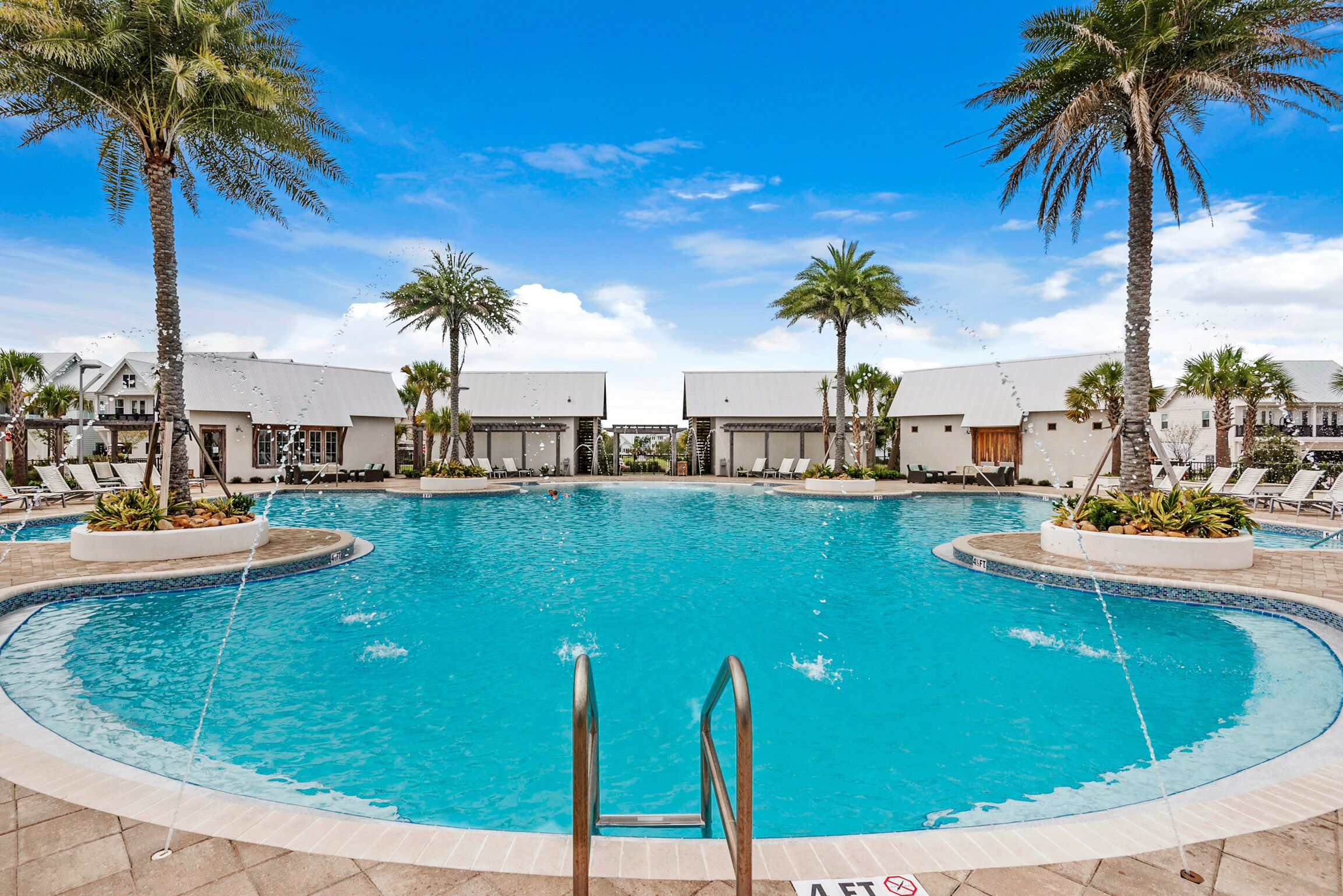 A large, clear swimming pool with palm trees and lounge chairs, surrounded by buildings with a modern design.