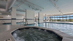 A large indoor swimming pool with a hot tub area, clear ceiling pipes, and a view of the outdoors through large windows.