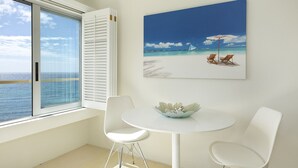 Une pièce avec une grande fenêtre, une table blanche et deux chaises, ainsi qu’un tableau représentant une scène de plage.