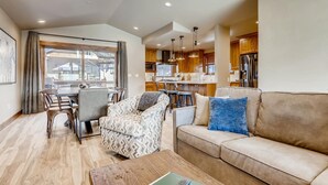Amplia sala de estar con sofá, sillón estampado y mesa de comedor. Se ve una cocina con armarios de madera y un ventanal grande al fondo.