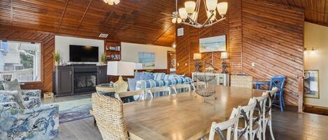 A spacious living room with a large wooden ceiling, a dining area with a long table and chairs, and a television mounted on the wall.