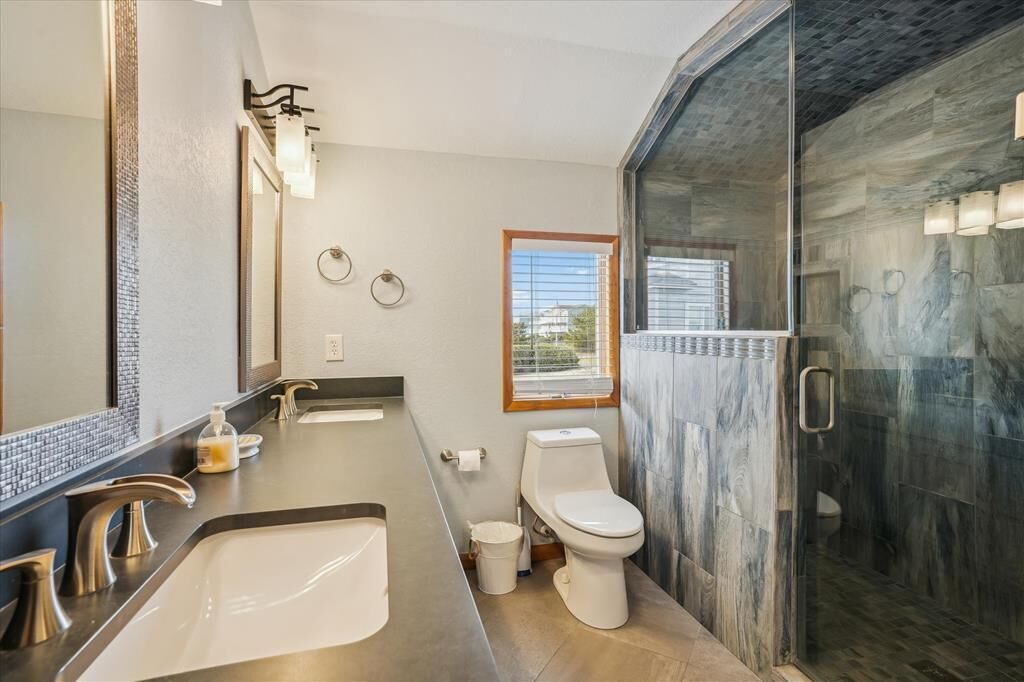 A modern bathroom with a large mirror, two sinks, a toilet, and a glass-enclosed shower area.