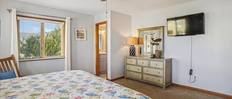 Unwind in your bright, airy bedroom with a cozy bed, ceiling fan, TV for entertainment, and a view of lush greenery through the window.