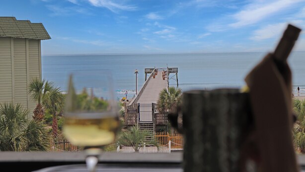 A view of a beach with a wooden boardwalk leading to the water, palm trees, and a building in the background. - Outstanding Oceanfront Condo with Private Balcony! (Isle of Palms)