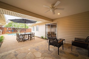 A covered patio with stone flooring, a table with chairs, a ceiling fan, and a pool in the background. - Best of Richardson | 5BD/3BA with Pool, Patio, BBQ (Richardson)