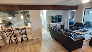 A modern living room with a black leather sofa, a coffee table, and a TV. There is a kitchen area with a fridge, stove, and bar stools. The room has wooden flooring and a large window.