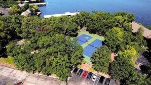 An aerial view of a tennis court surrounded by dense greenery, with a parking area in the foreground.