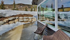Une terrasse fermée par des baies vitrées, avec un mur de soutènement en pierre, une chaise et une vue sur un paysage enneigé, avec des arbres et une montagne.