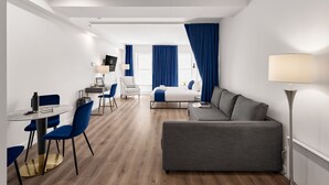 Een moderne hotelkamer met een grijze bank, een klein rond tafeltje met blauwe stoelen, een bed met blauwe gordijnen en een bureau met stoel.