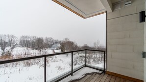 Un balcón con piso de madera y baranda de vidrio, con vista a un paisaje nevado con casas y árboles desnudos.