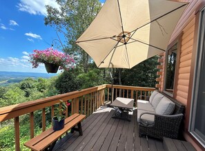 A wooden deck with a beige umbrella, wicker furniture, and hanging flower baskets. - Cozy Mountaintop Retreat (New Paris)