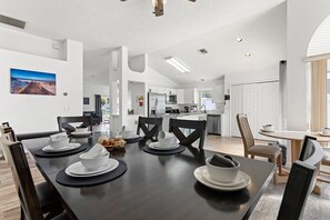 Un coin repas moderne avec une table en bois foncé dressée pour six personnes, des assiettes blanches et des sets de table noirs. On aperçoit une cuisine en arrière-plan, avec des armoires blanches et des appareils en acier inoxydable.