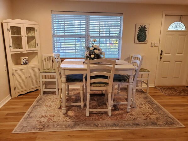 Un comedor con mesa y sillas, una ventana con persianas, un armario con puertas de vidrio y una puerta con un panel de vidrio decorativo.
