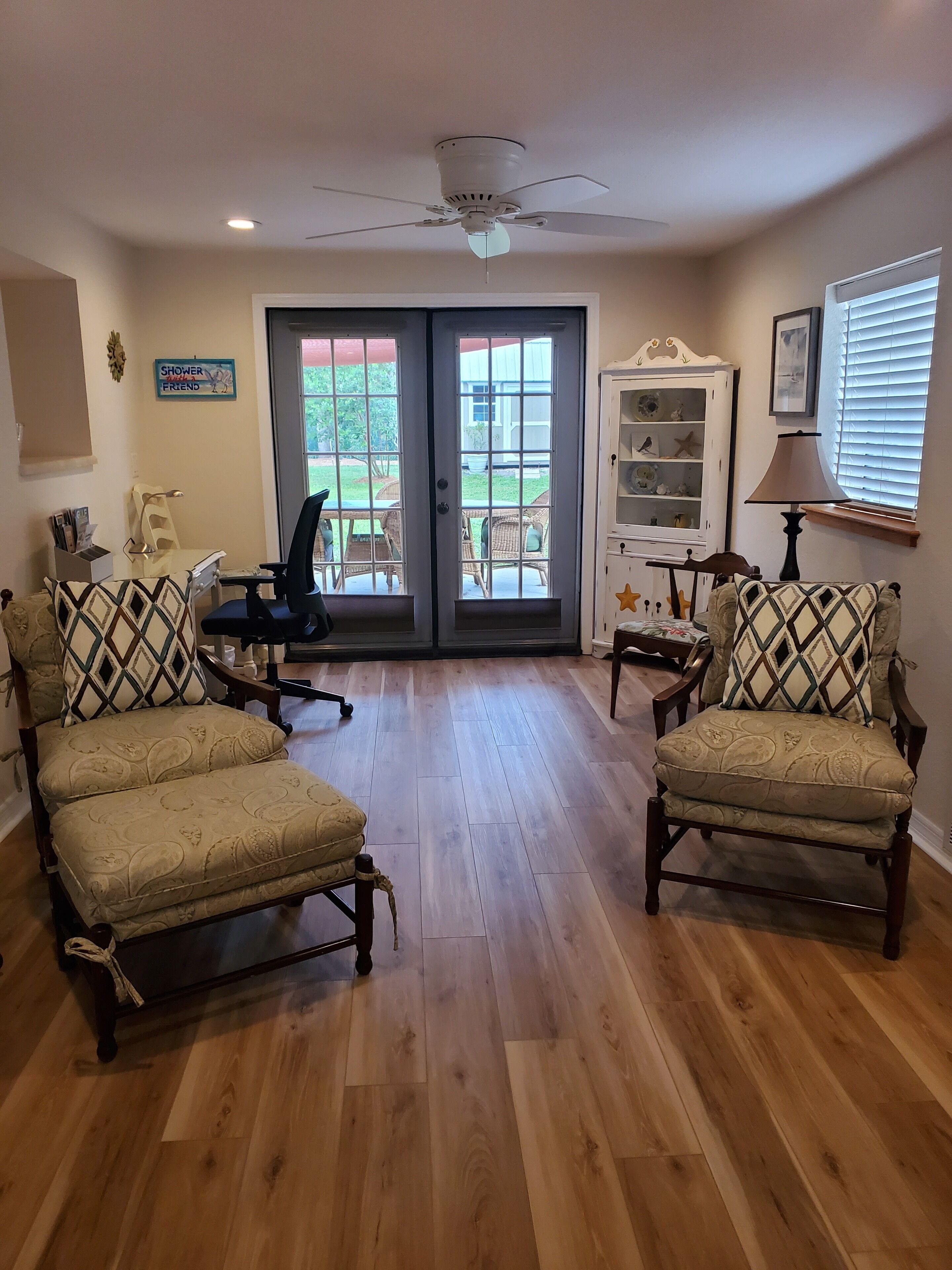 A spacious room with wooden flooring, two armchairs, a desk, a chair, a ceiling fan, and a view through a glass door to an outdoor patio.