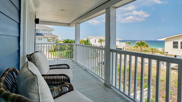 A covered porch with white railings, wicker furniture, and a view of the ocean and beach houses. - Experience island life with amazing views and easy beach access! 6BR, 5BA (Wrightsville Beach)