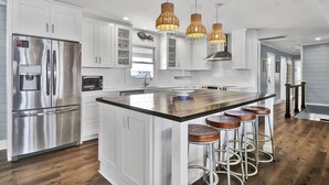 A modern kitchen with white cabinetry, stainless steel appliances, a central island with stools, and three wicker pendant lights. - Experience island life with amazing views and easy beach access! 6BR, 5BA (Wrightsville Beach)