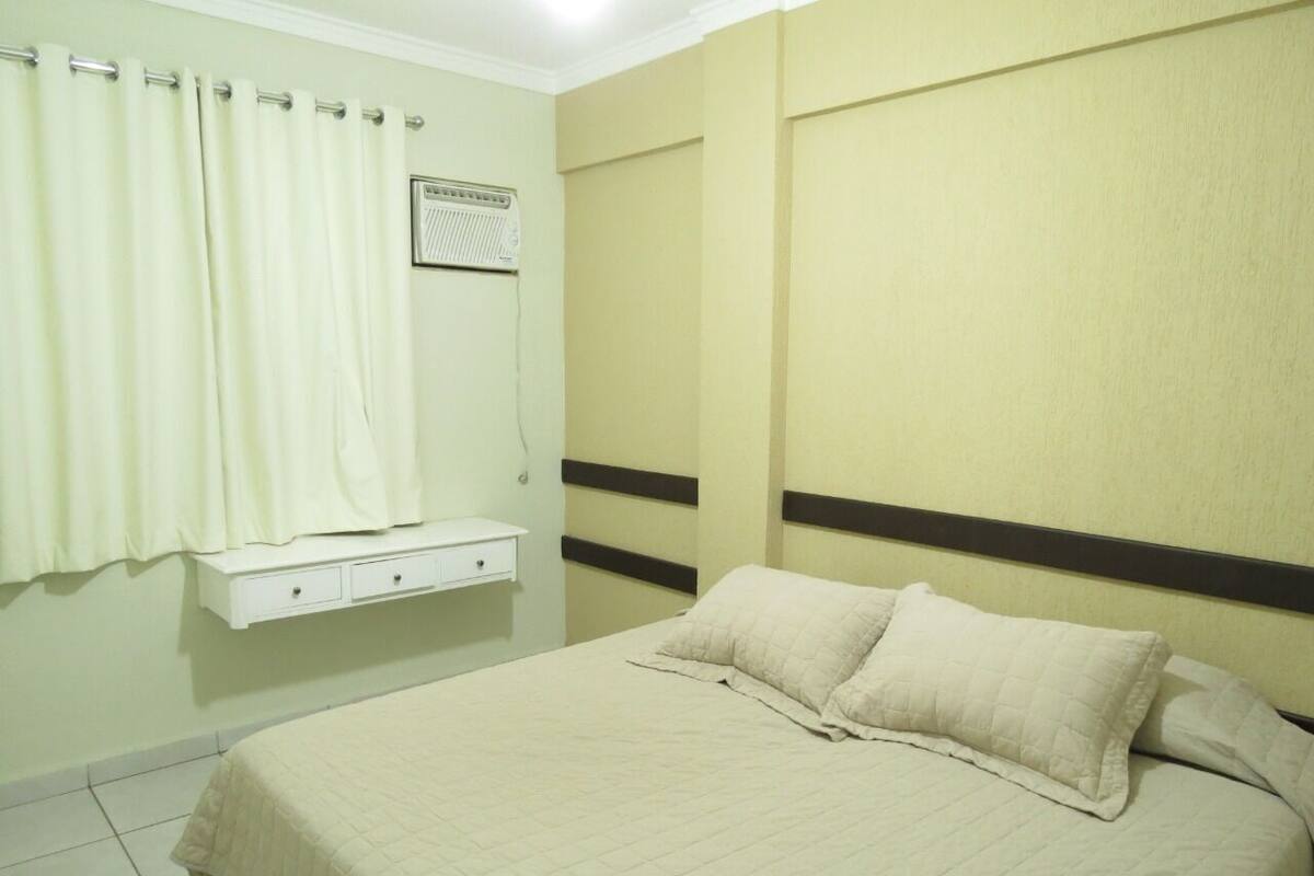 A bedroom with a bed, a bedside table, a wall-mounted heater, and a window with curtains.