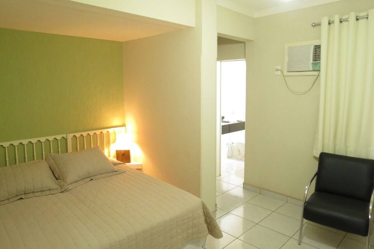 A hotel room with a bed, a chair, a nightstand with a lamp, and an air conditioning unit on the wall.