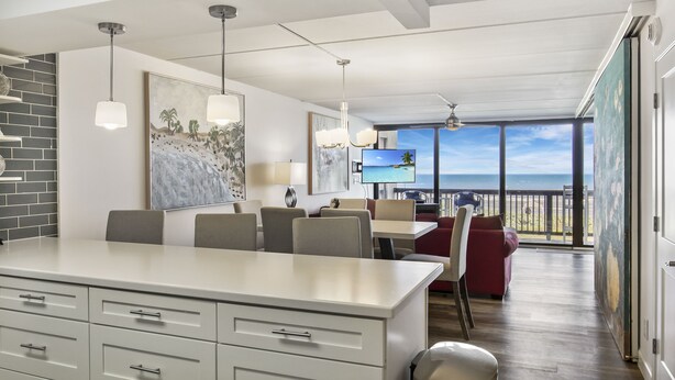 A modern dining area with a white island, grey chairs, and a red sofa. There are pendant lights and a TV on a wall.