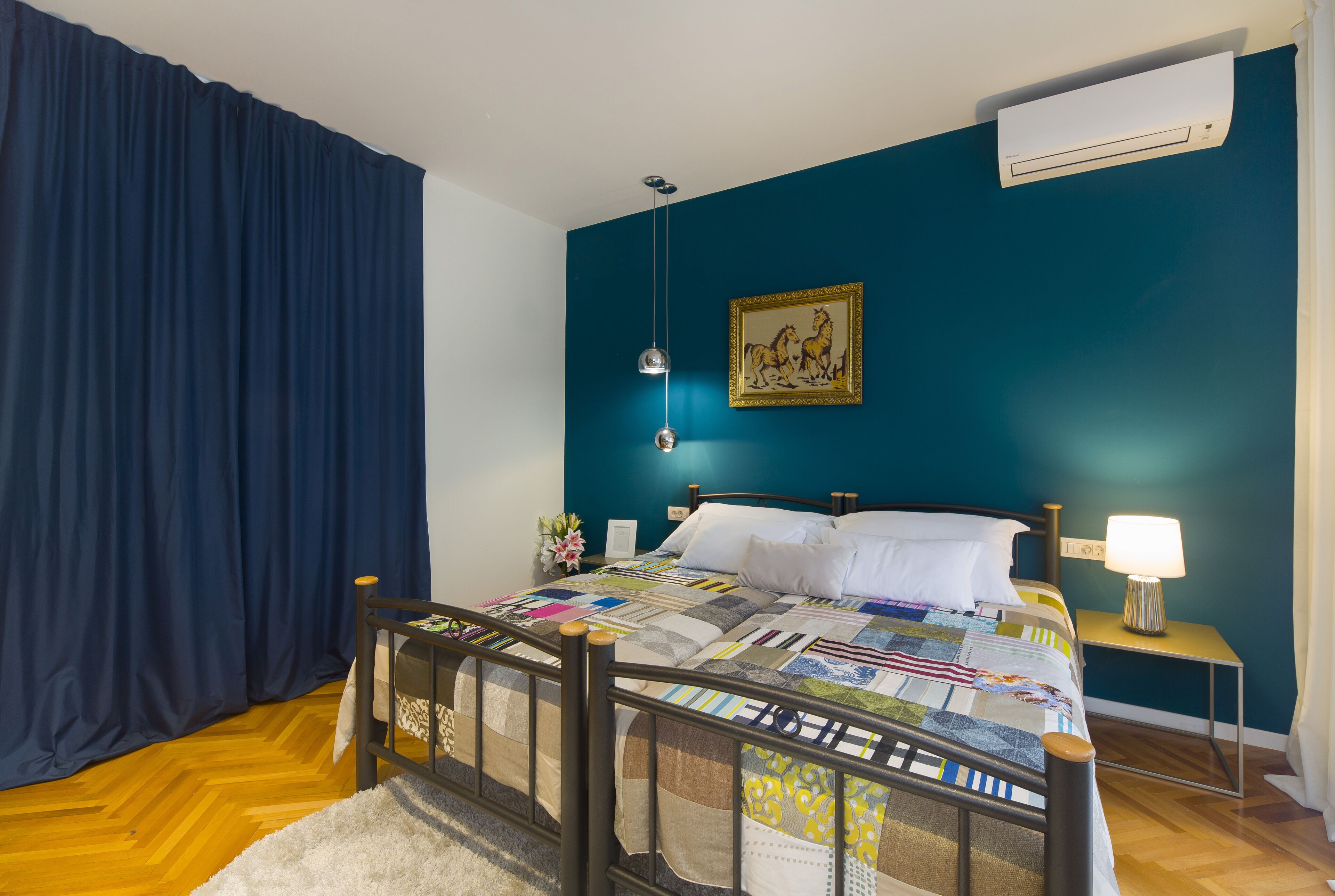 A bedroom with a wooden bed, a blue wall, an air conditioner, a bedside lamp, and a framed picture on the wall.