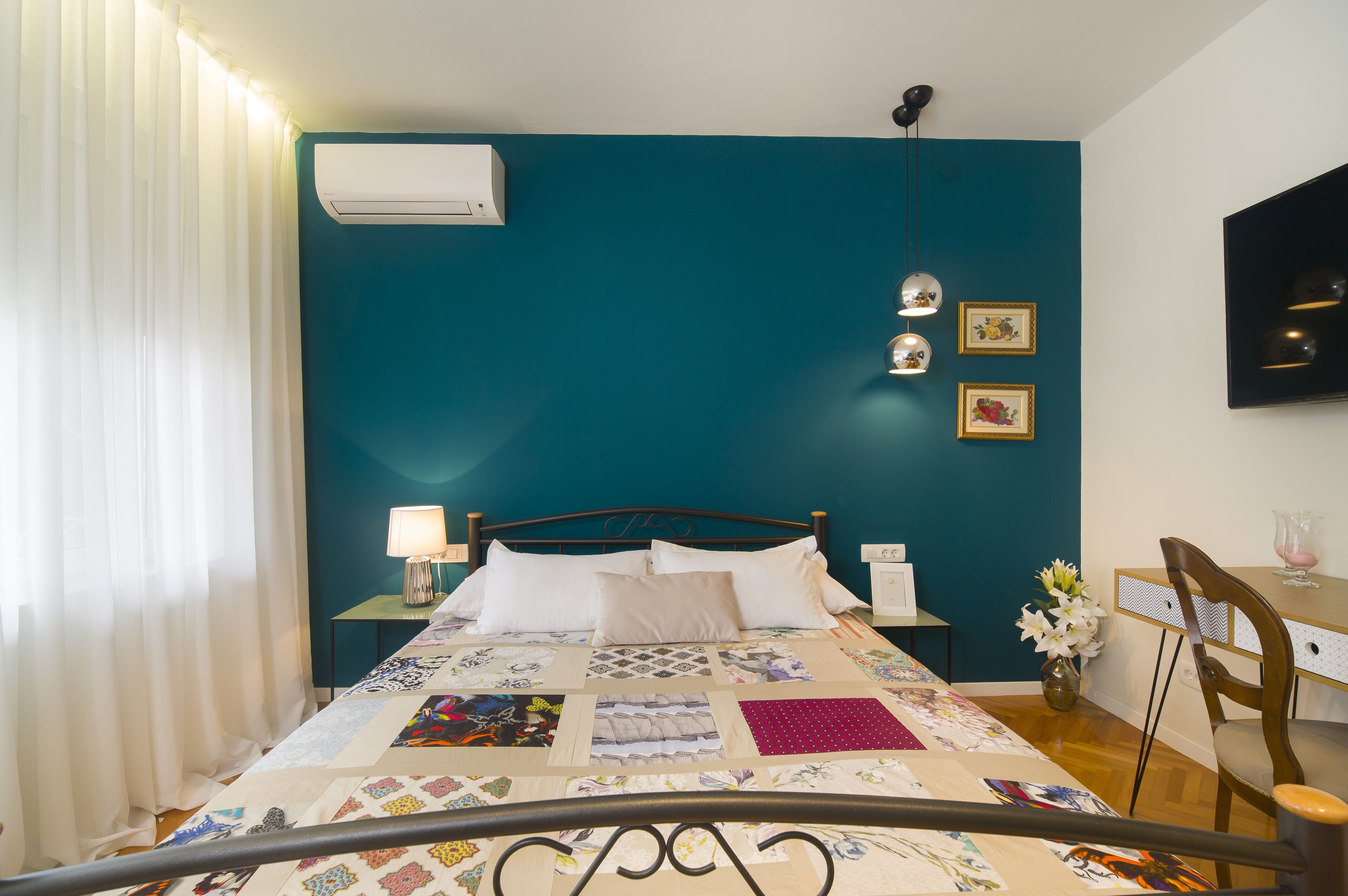 A bedroom with a teal wall, a bed with a quilt, a bedside table with a lamp, a desk with a chair, and a wall-mounted TV.