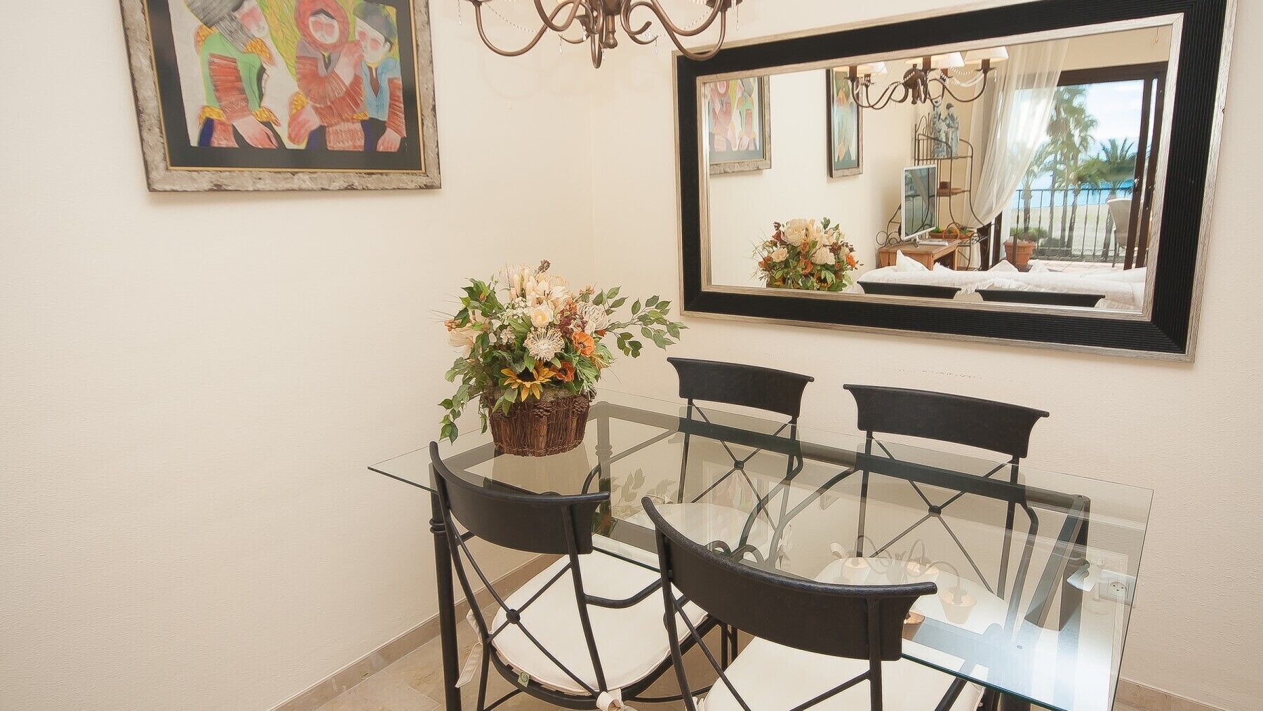 A dining area with a glass table and black chairs, a potted plant, and a large mirror reflecting a beach view.