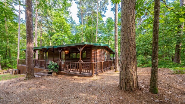 A rustic cabin with a porch and a small deck, surrounded by tall trees in a wooded area.