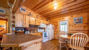 A wooden cabin interior with a kitchen area featuring a stove, microwave, refrigerator, and wooden cabinets. There is a dining table with chairs and a window with a view.