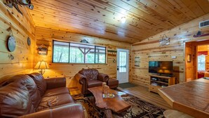 A cozy cabin interior with a leather sofa, a wooden coffee table, a TV, and a large window.