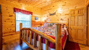 A wooden cabin bedroom with a bed, a window with a red valance, and a door.