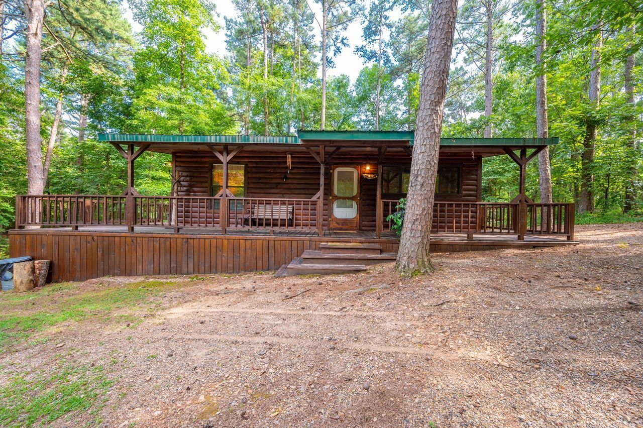 Una cabaña rústica con un porche cubierto y una pequeña terraza, rodeada de un bosque denso.
