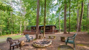 A cabin surrounded by tall trees, with outdoor seating and a fire pit.