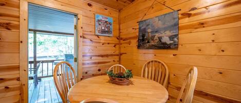 Interior de una cabaña de madera con una mesa redonda, cuatro sillas de madera, una cesta con plantas, un cuadro en la pared y una puerta que da a una terraza exterior.