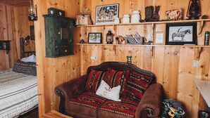 Interior acogedor de una cabaña con techo de madera, un sofá de cuero marrón, un tapiz de piel de vaca y estantes con diversos objetos, incluyendo un cuadro enmarcado de un caballo.