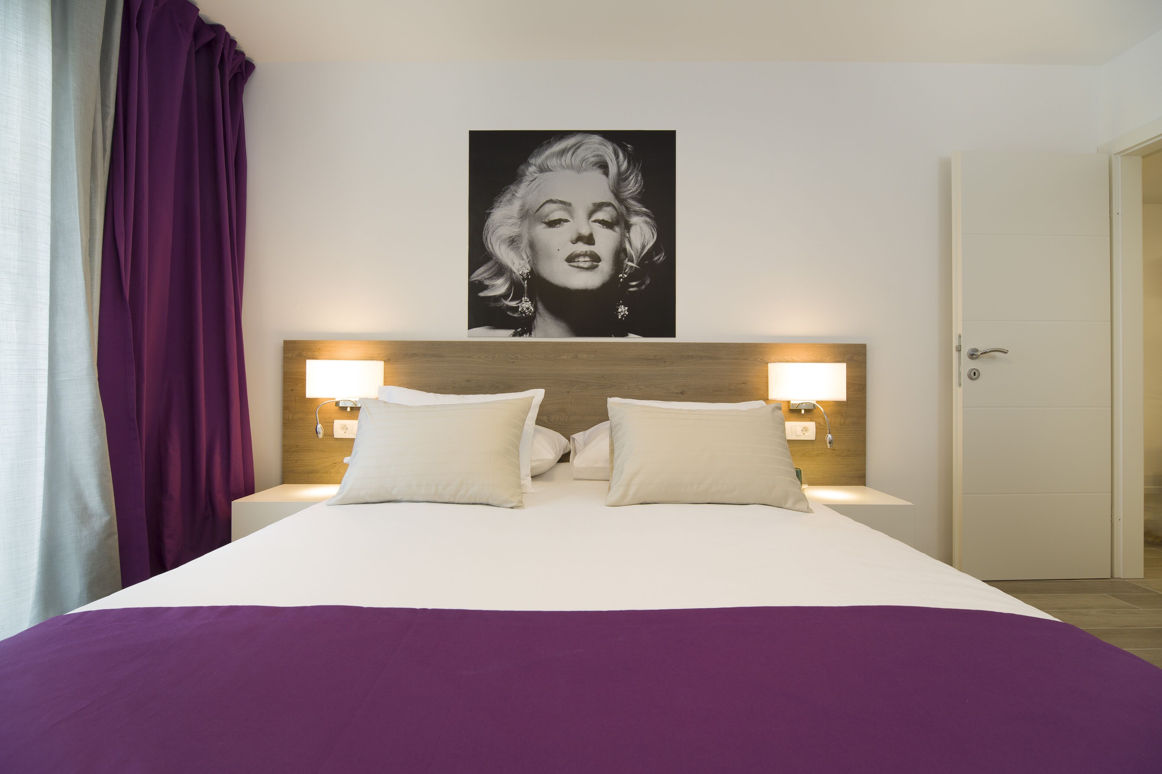 A hotel room with a large bed, two bedside lamps, a wooden headboard, and a poster on the wall.
