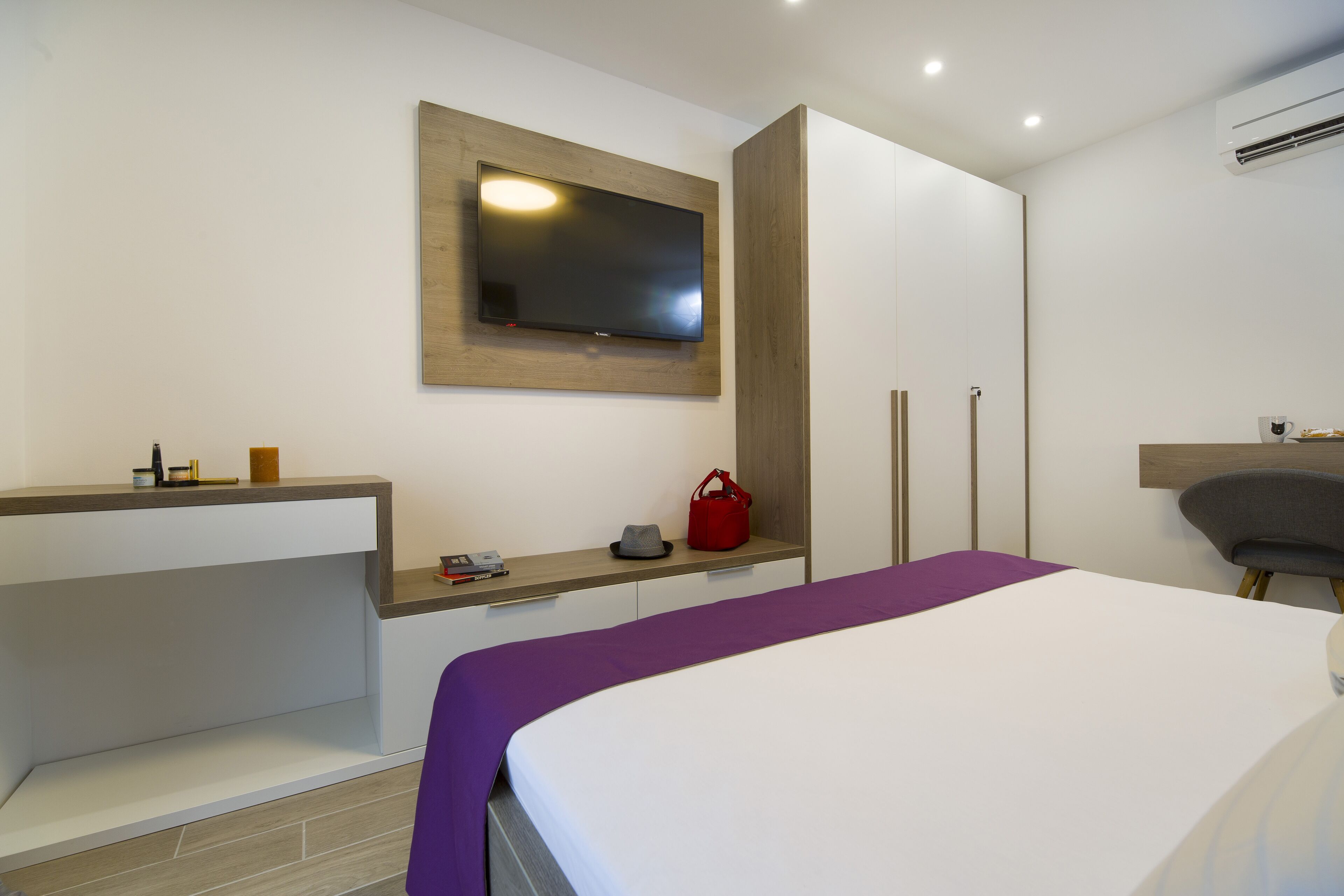 A modern hotel room with a bed, a TV mounted on the wall, a vanity with a mirror, and a small seating area.
