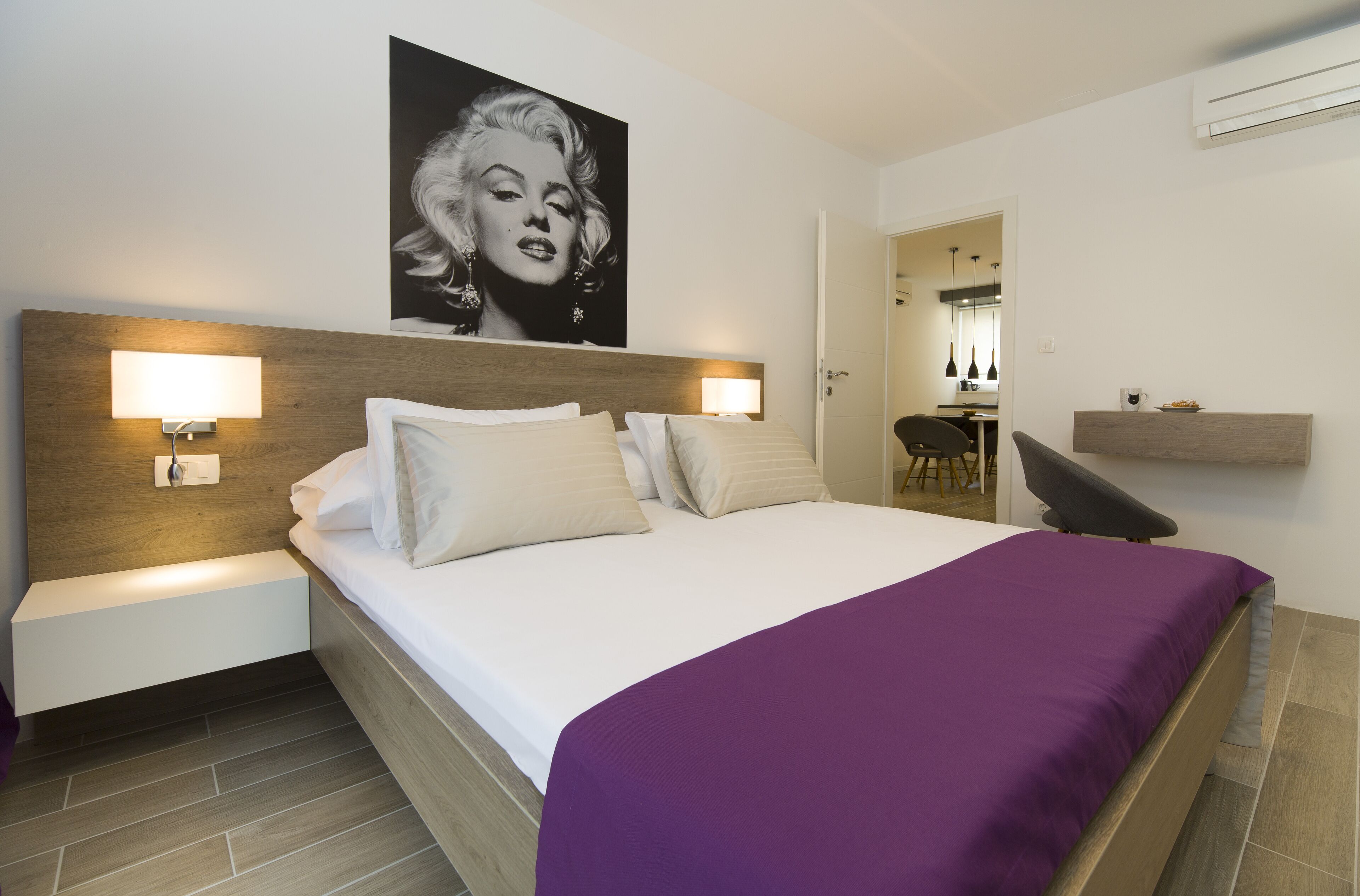 A hotel room with a bed, two bedside lamps, a wooden headboard, a purple bedspread, a wall-mounted picture, and a small desk with a chair.