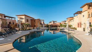 Una piscina cristalina con sillas de descanso y una pérgola, rodeada de edificios de estilo mediterráneo.