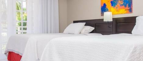 A hotel room with two beds, white bedding, a red skirted bed base, a large abstract painting on the wall, and a window with white curtains.