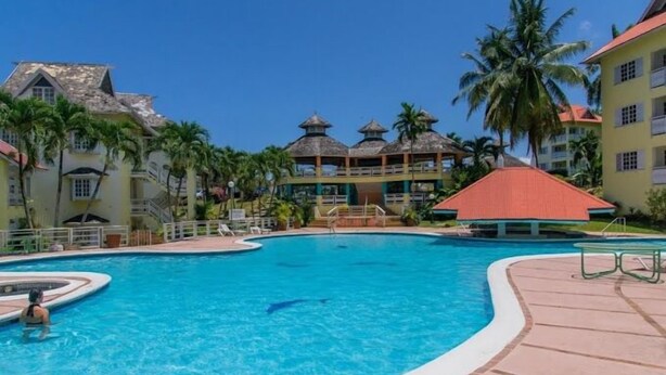 A resort pool area with a large central pool, palm trees, and resort buildings with red roofs. - Mystic Ridge Resort (Ocho Rios)