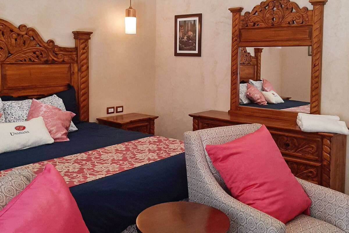 A hotel room with a large bed, a sitting area with a sofa and armchairs, a small round table, a wooden headboard, a wall-mounted mirror, and a ceiling light fixture.