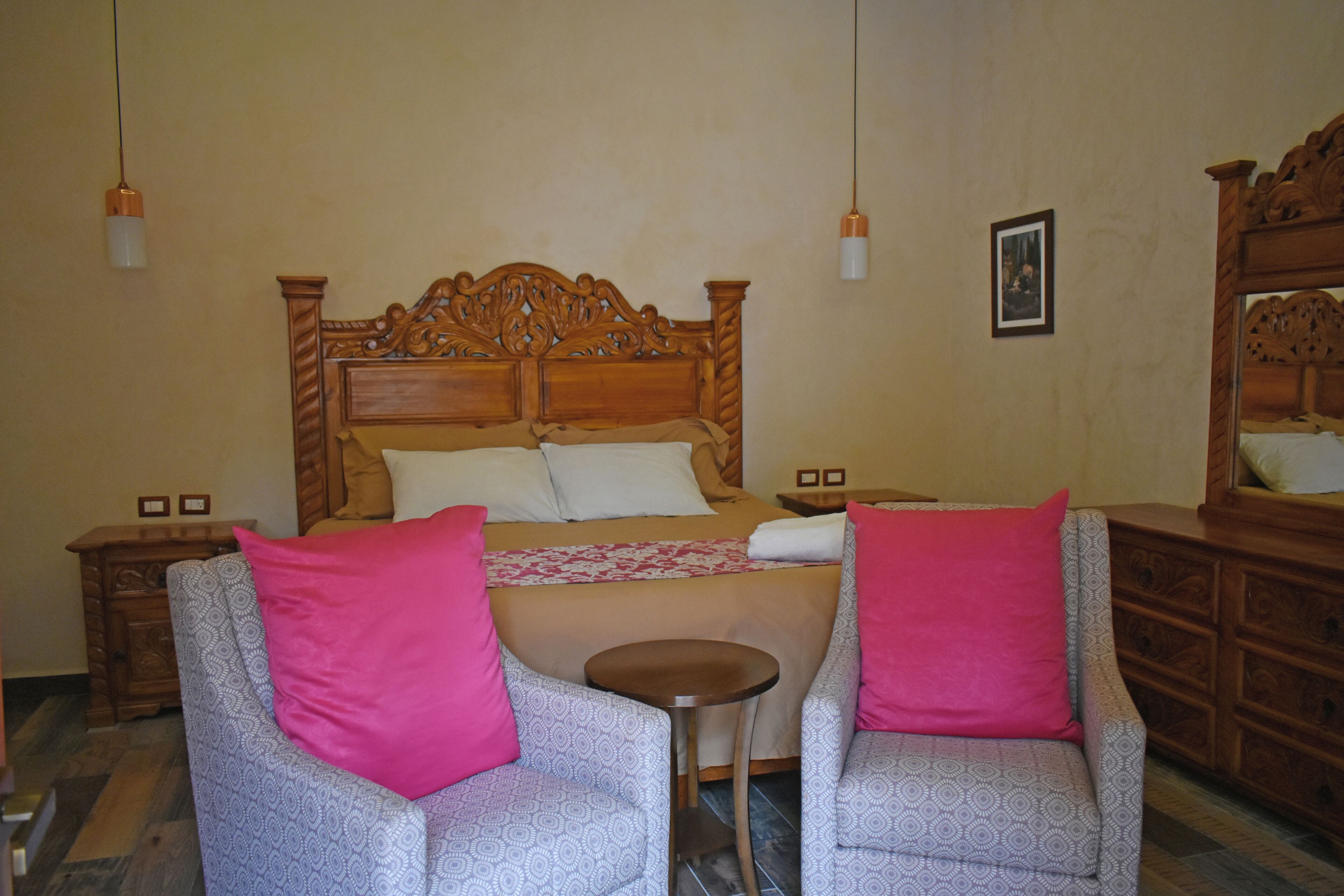 A bedroom with a wooden bed, two chairs with pink cushions, a small round table, and a dresser with a mirror.