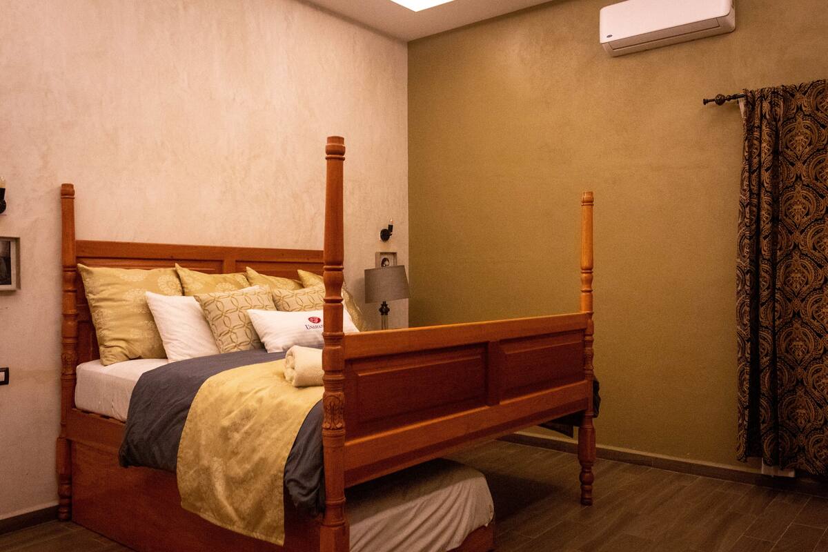 A four-poster bed with a wooden frame and a headboard, a bedside table with a lamp, and a patterned curtain on a window.