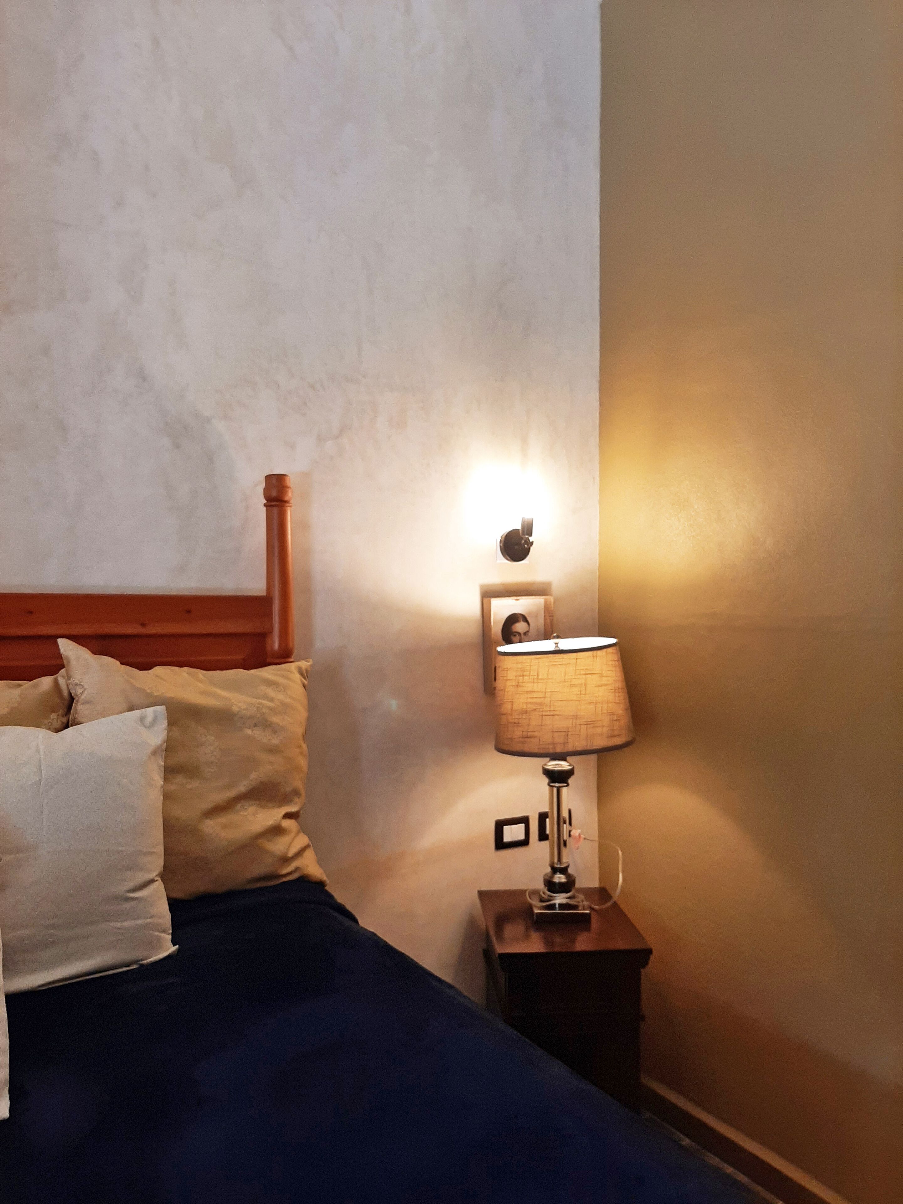 Una cama con cabecera de madera, una mesita de noche con lámpara y una luz empotrada en la pared.