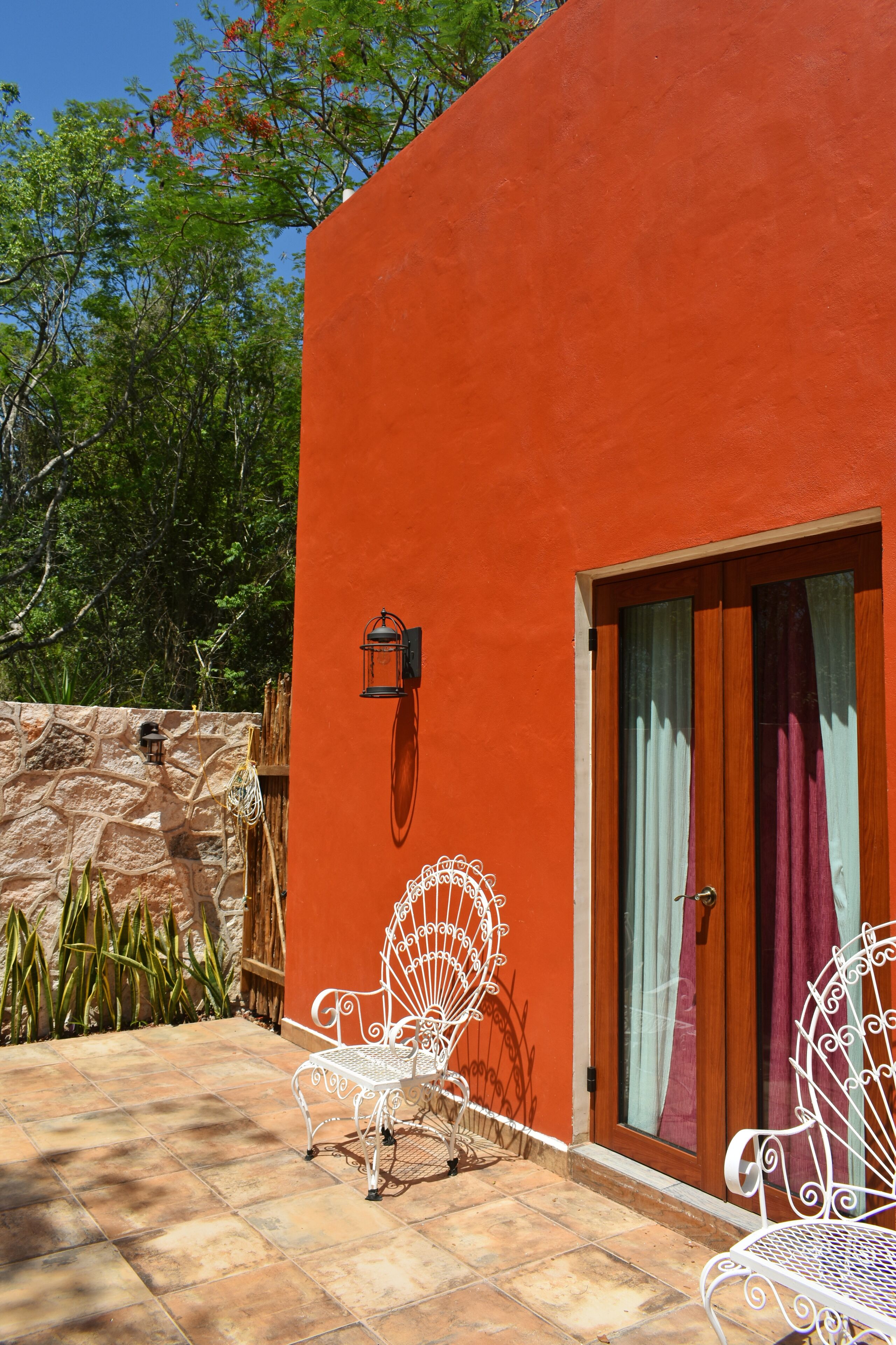 Un patio con dos sillas de metal blanco, un muro de piedra y una puerta de madera con cortinas.