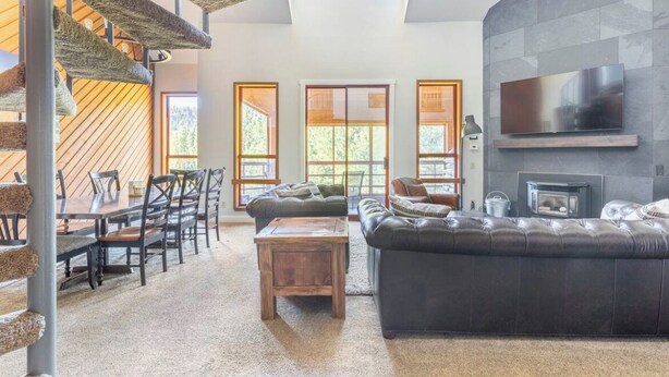 A spacious living room with a high ceiling, a fireplace, a large TV mounted on the wall, and a staircase with a unique design. - Aspen 230-Walking distance to the ski area with views of Mammoth Mountain! (Mammoth Lakes)