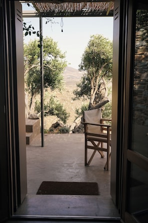 An open doorway leading to a patio area with a view of a mountainous landscape, a wooden chair, and a stone wall. - Avdos Olive House · off-grid Cycladic retreat (Tinos)