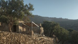 A stone building with a sloped roof, surrounded by olive trees and a mountainous backdrop. - A house in an olive grove with a unique view to the Aegean sea. (Tinos)