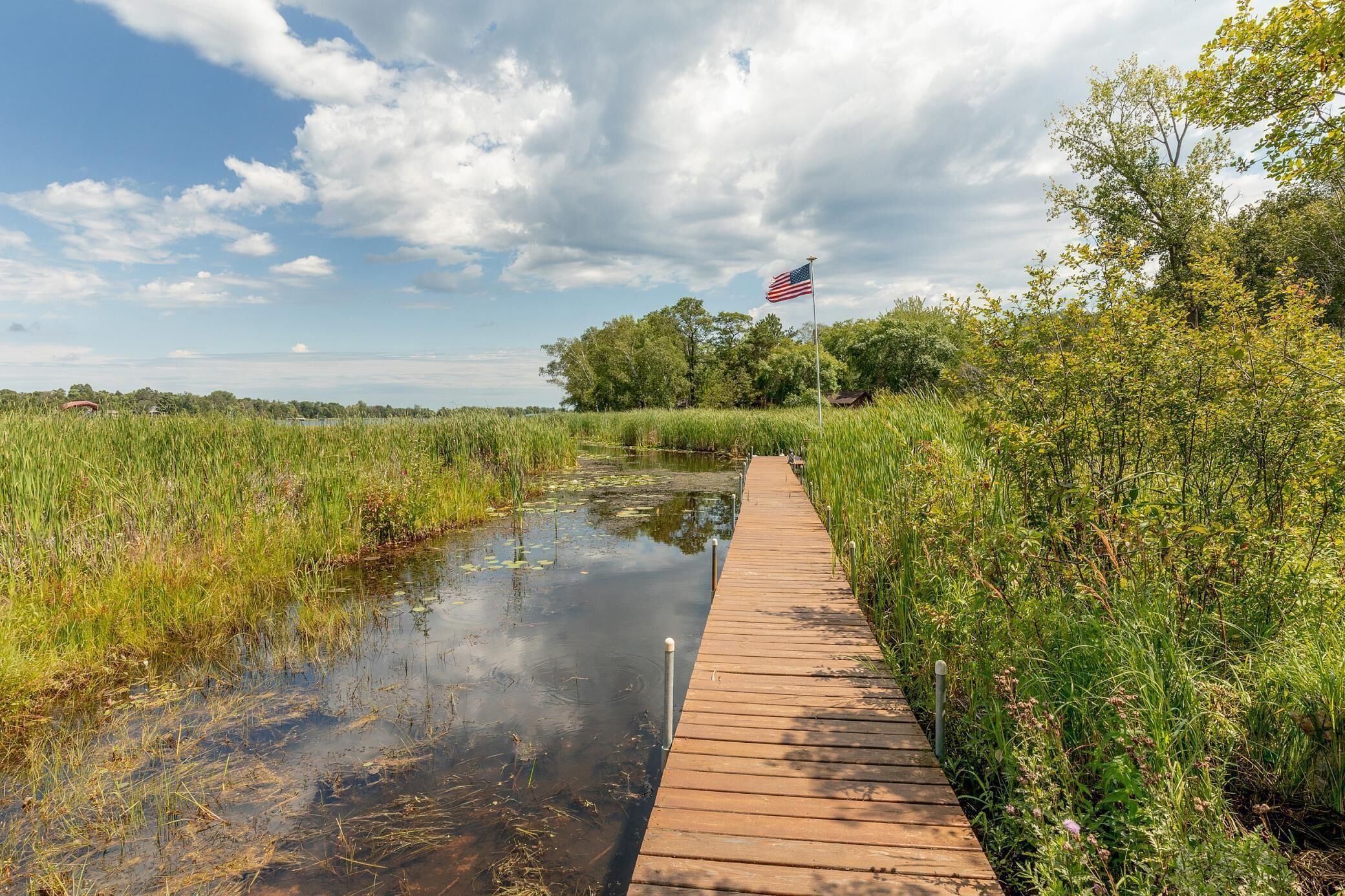 A wooden boardwalk beside a calm water body with an American flag, surrounded by lush greenery and trees.