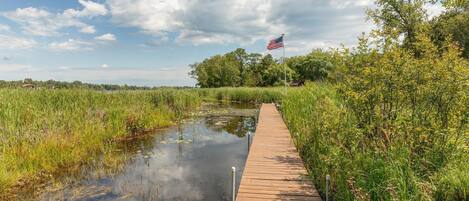 A wooden boardwalk beside a calm water body with an American flag, surrounded by lush greenery and trees.